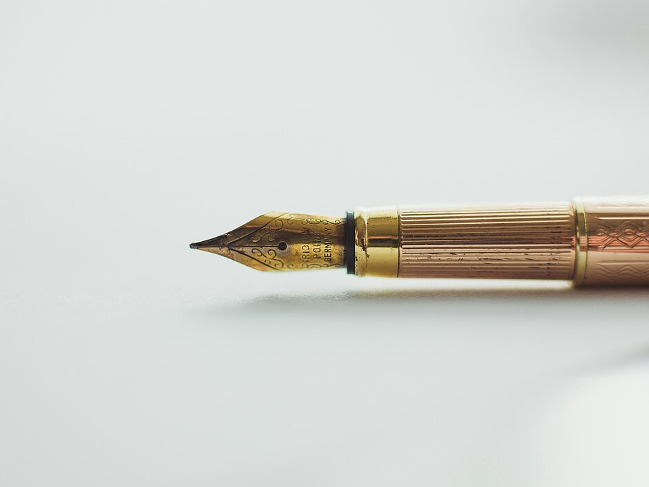 Nahaufnahme einer goldfarbenen Feder eines klassischen Füllfederhalters auf hellem Untergrund.