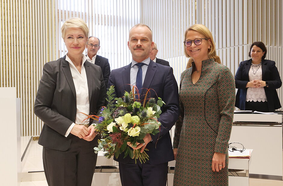 Ein Mann im dunklem Anzug und eine Frau im hellen Kostüm reichen sich im Plenarsaal des Landtags Mecklenburg-Vorpommern die Hand, umgeben von stehenden Abgeordneten mit Blumensträußen - anlässlich einer Wahl zum Bürgerbeauftragten