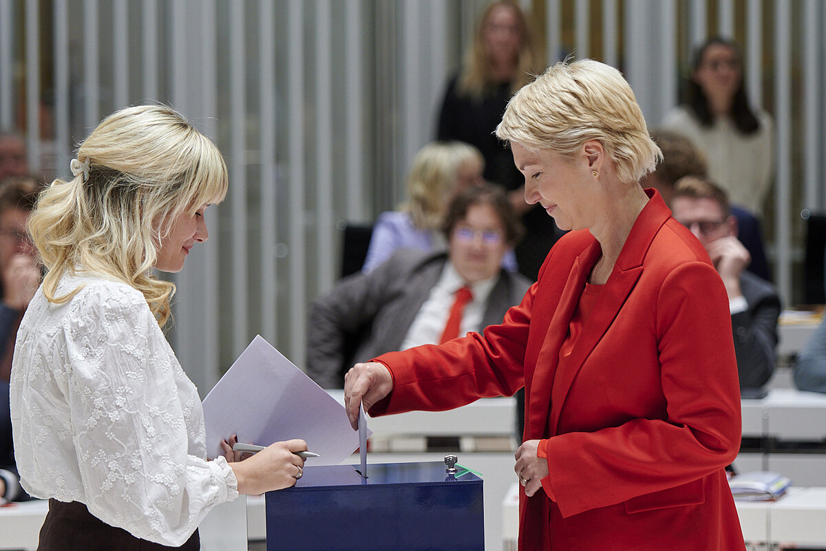 Frau in rotem Kostüm wirft Wahlzettel in die Urne