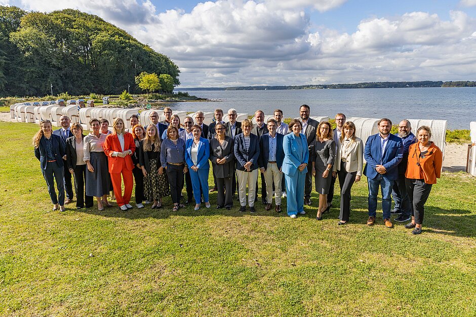 Gruppenfoto der Teilnehmenden der 20. Parlamentarischen Ostsee-Konferenz (PSO) 2024 in Glücksburg, aufgenommen bei sonnigem Wetter auf einer Wiese vor dem Strand mit Strandkörben und Blick auf die Ostsee.