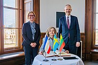 Drei Personen, zwei Frauen und ein Mann in einem repräsentativen Raum, auf dem Tisch vor ihnen sind die Flaggen von Deutschland, Schweden und Litauen platziert.