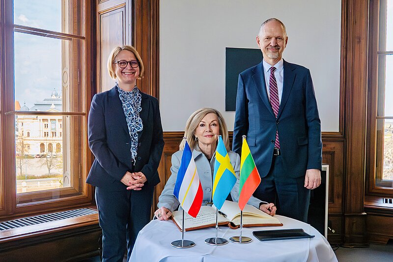 Drei Personen, zwei Frauen und ein Mann in einem repräsentativen Raum, auf dem Tisch vor ihnen sind die Flaggen von Deutschland, Schweden und Litauen platziert.