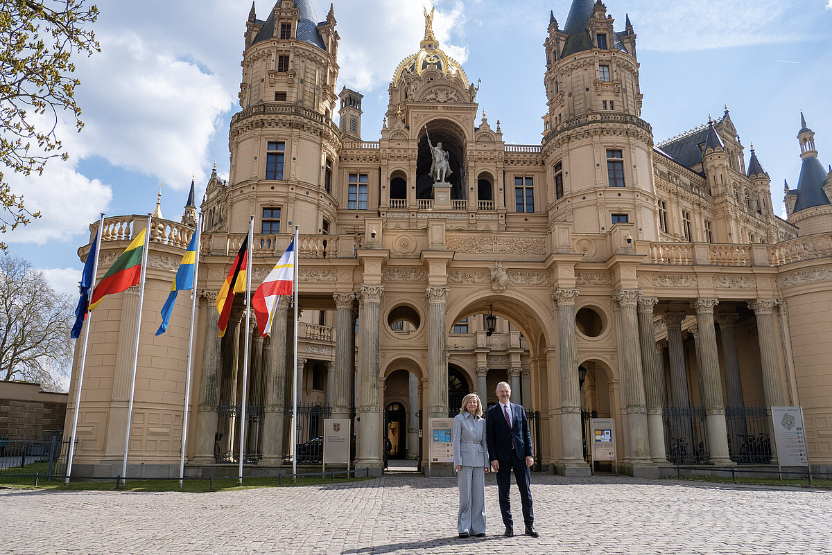 Ein Mann und eine Frau in Business-Anzügen stehen vor einem historischen Schloss an einem sonnigen Tag, es wehen die Flaggen von Deutschland, Litauen, Schweden, MV und Europa 