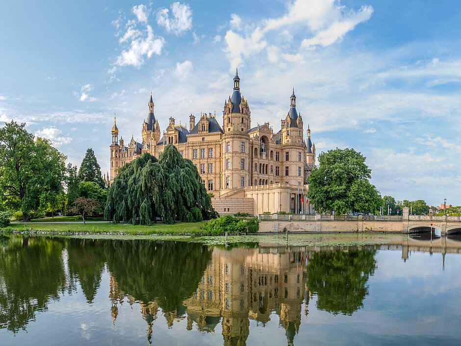 Panoramablick auf das Schweriner Schloss mit Türmen und reich verzierter Fassade, umgeben von Bäumen und Wasser, im Vordergrund eine steinerne Brücke über den Burgsee.