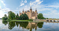 Panoramablick auf das Schweriner Schloss mit Türmen und reich verzierter Fassade, umgeben von Bäumen und Wasser, im Vordergrund eine steinerne Brücke über den Burgsee.