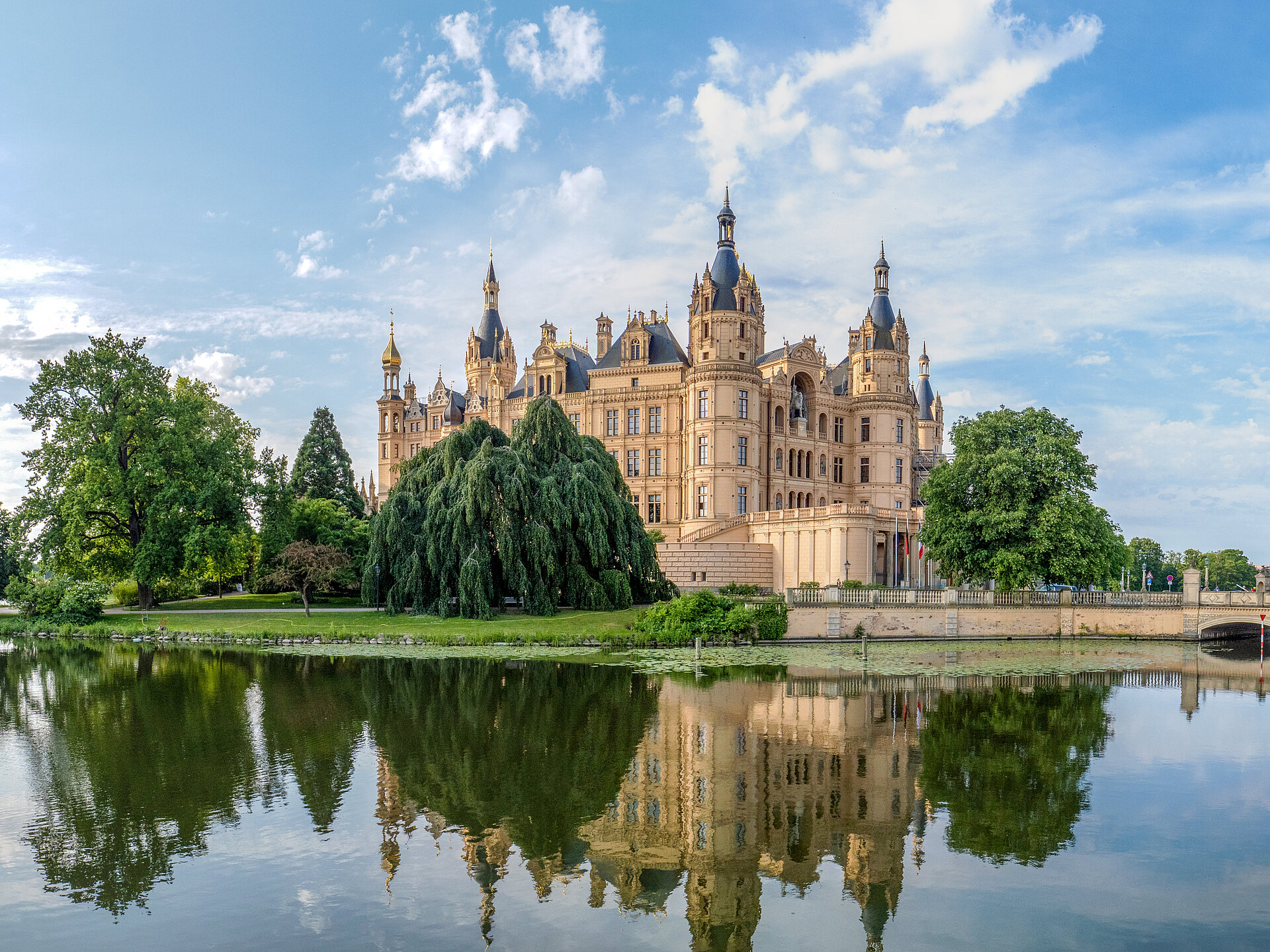 Panoramablick auf das Schweriner Schloss mit Türmen und reich verzierter Fassade, umgeben von Bäumen und Wasser, im Vordergrund eine steinerne Brücke über den Burgsee.