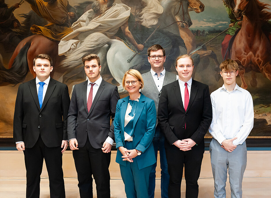 Fünf junge Männer und eine Frau in einem türkisfarbenen Anzug, stehen vor einem großformatigen historischen Gemälde im Schweriner Schloss und blicken in die Kamera.