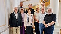 Gruppe von acht Personen steht vor einer Treppe in einem hellen Raum