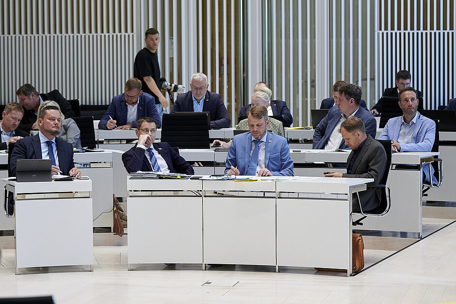 Abgeordnete im Plenarsaal des Landtags Mecklenburg-Vorpommern, mehrere Männer in Anzügen sitzen an weißen Tischen und machen sich Notizen