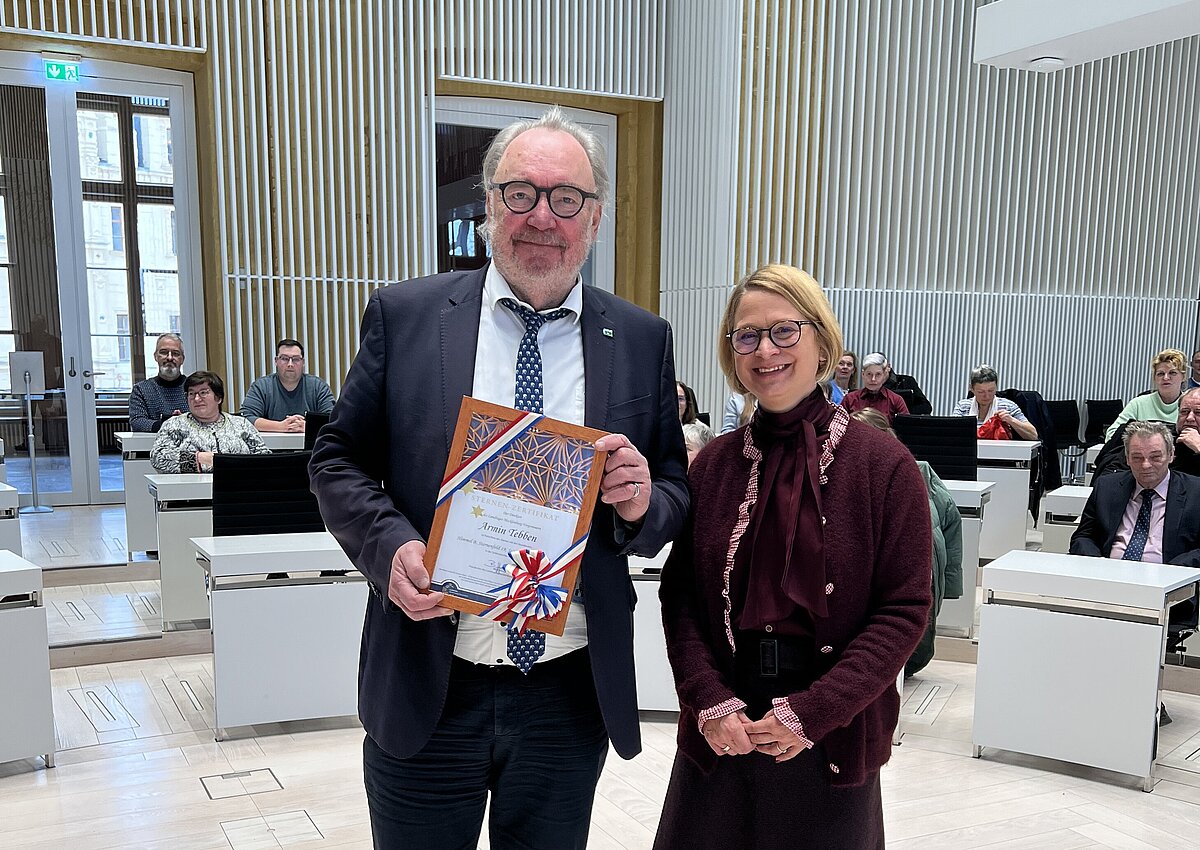 Ein Mann hält eine Urkunde in der Hand, eine Frau steht neben ihm im Plenarsaal