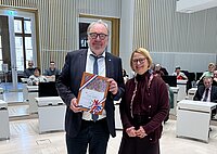 Ein Mann hält eine Urkunde in der Hand, eine Frau steht neben ihm im Plenarsaal