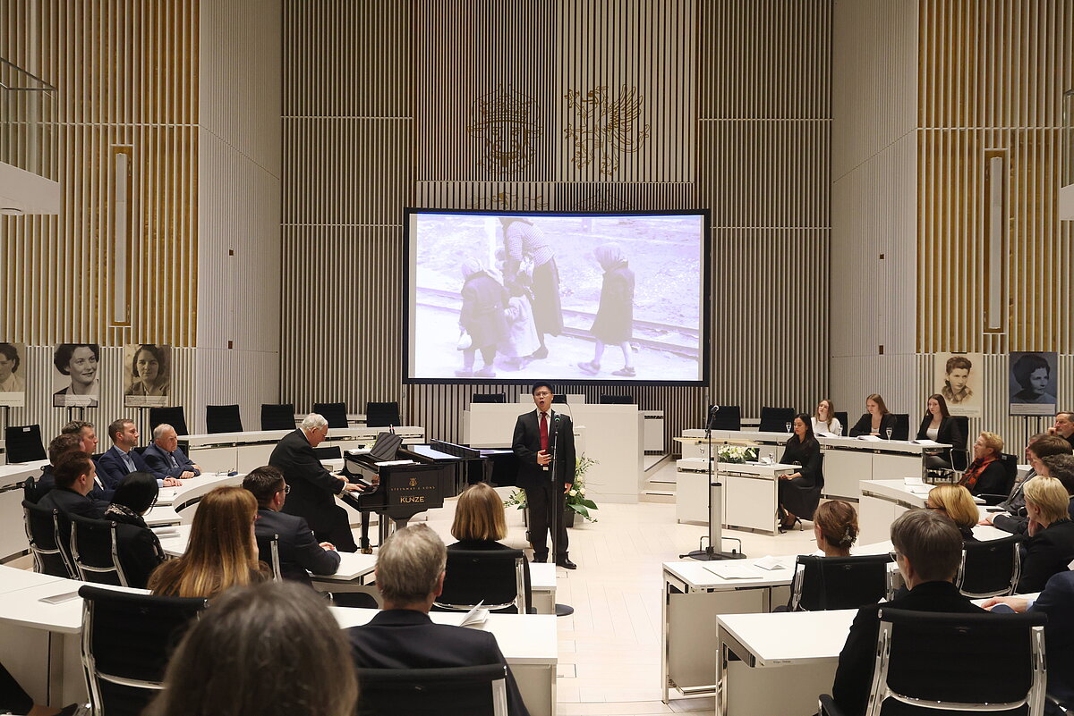 Blick in den Plenarsaal mit anwesendem Publikum; ein Sänger steht in der Mitte des Saals, im Hintergrund eine Leinwand mit Bildprojektionen